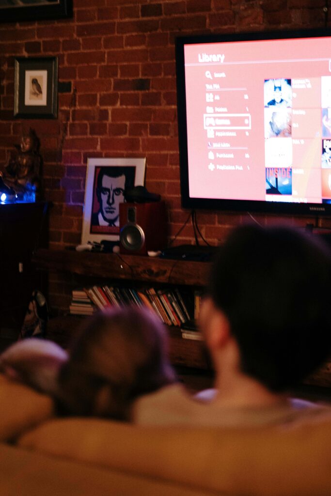 A man and his partner watching TV together, with a music speaker, books, and an art painting in the background.