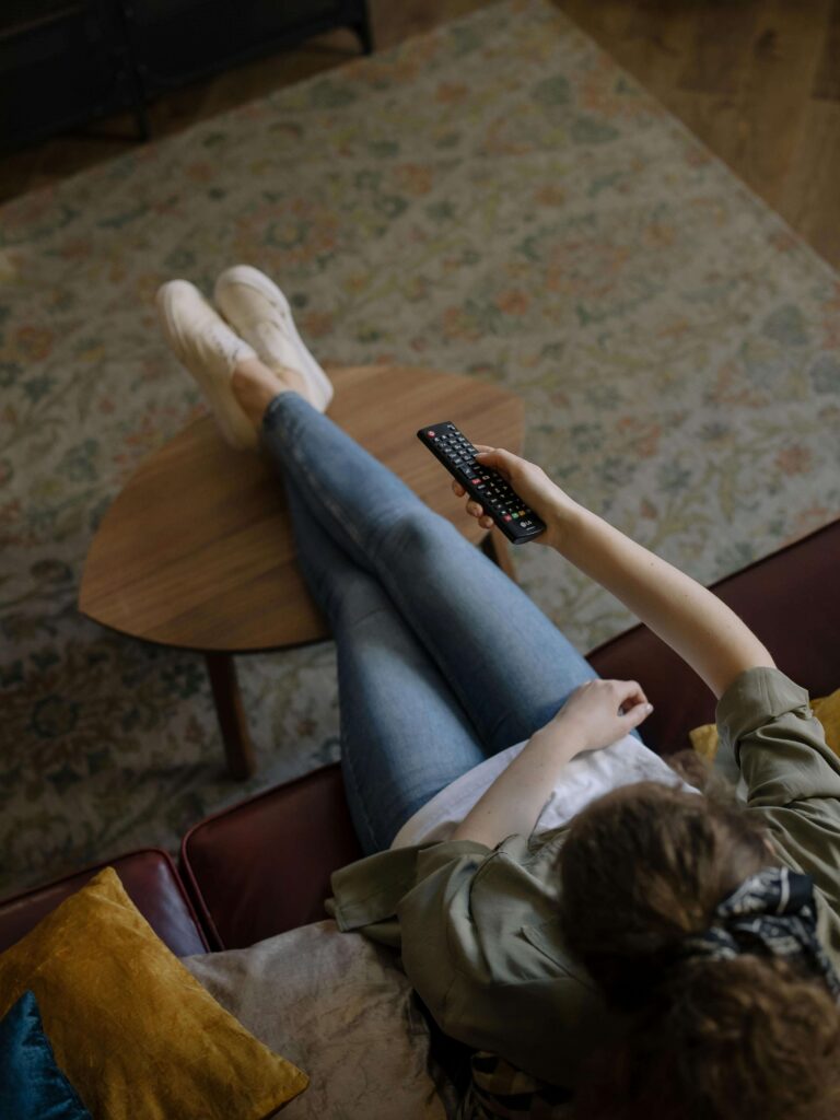 Woman sitting on a sofa with her legs up on a dining table, switching through TV channels using a remote control.