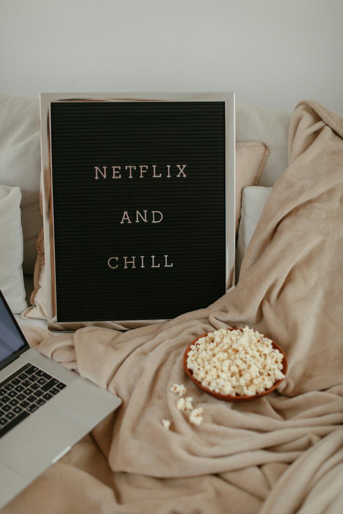 Blackboard sign with ‘Netflix and Chill’ written on it, next to a laptop and a box of popcorn — representing a cozy movie night setup.