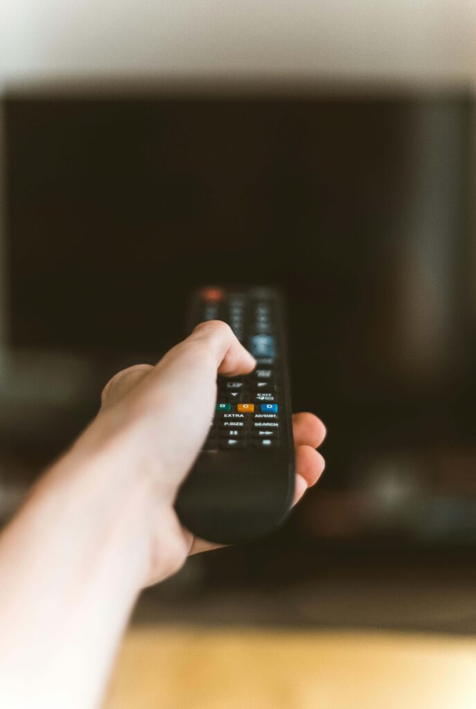 A hand reaching out to power on the television using a remote control — a moment just before the screen lights up.