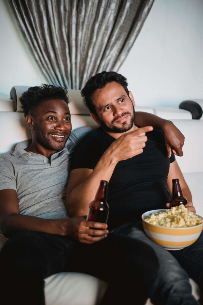 Two friends enjoying a movie together, holding a bottle of wine and a bowl of popcorn in a cozy, relaxed setting.