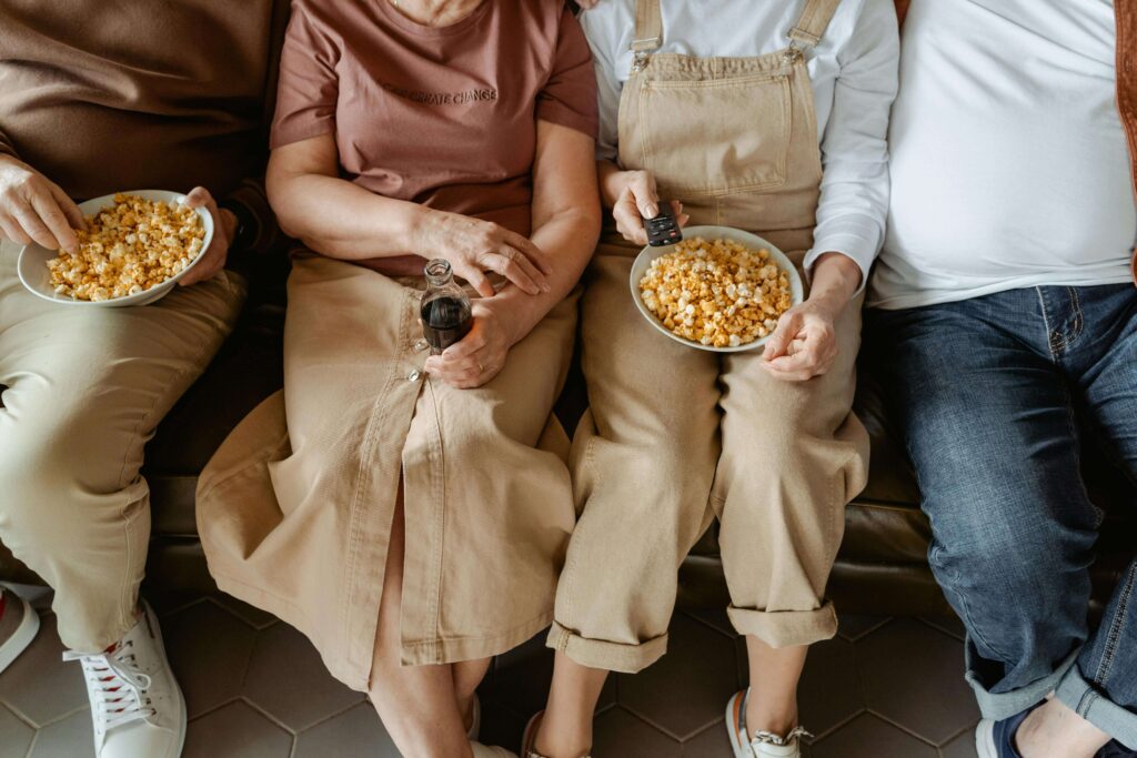 A smiling family of four—two parents and two kids—sharing a bowl of popcorn and watching a movie on TV in a warm, comfortable living room.