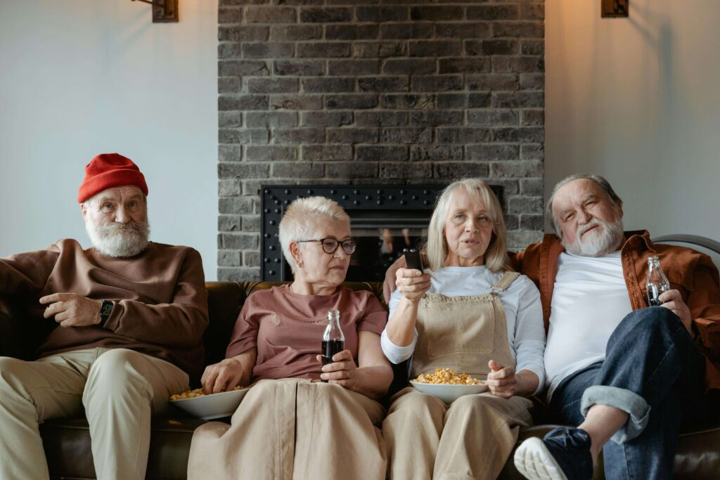 Elderly family group with two women and two men enjoying TV together, sharing snacks and drinks in a cozy living room.