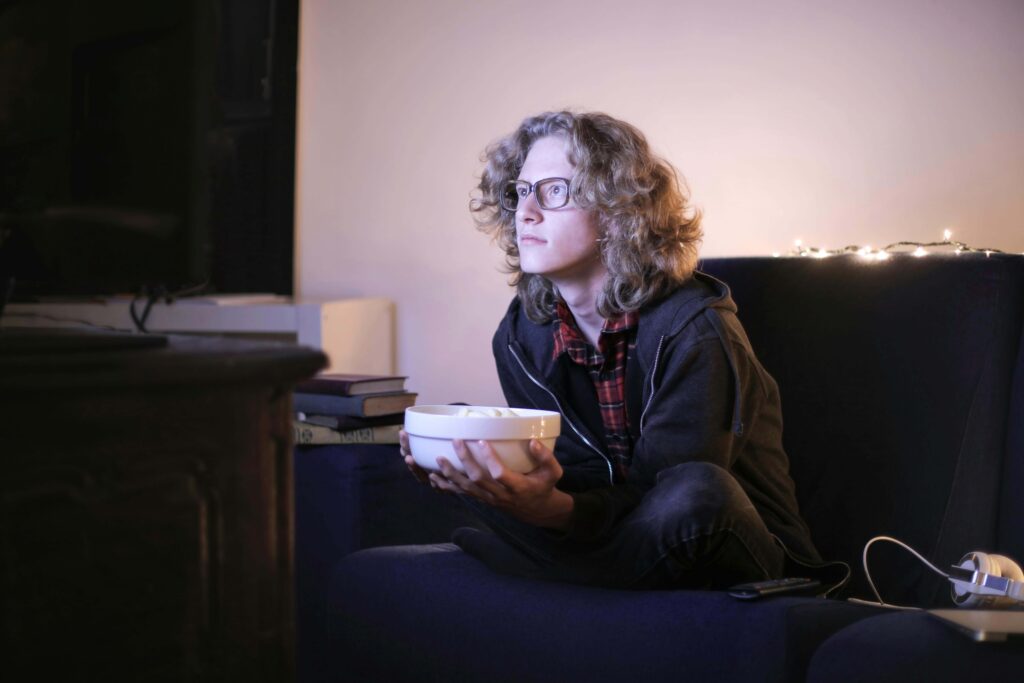 A woman is sitting cross-legged while watching the large TV screen in front of her. She holds a plate filled with her favorite food in her hands, picking up a bite with a calm smile that shows she’s enjoying the moment. The soft light from the screen gently illuminates her face, creating a warm and cozy atmosphere in the room.