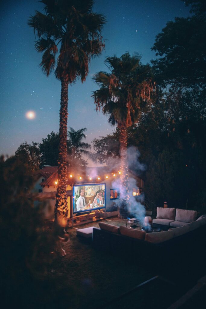 An outdoor setup featuring a TV screen displaying a scene, with a cozy couch placed in the open air under the evening sky.
