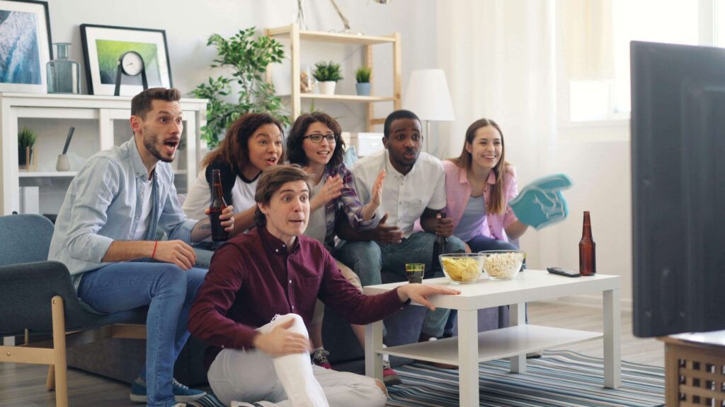 Six friends from diverse backgrounds — three women and three men — happily enjoying a TV show together. A bowl of popcorn sits on the table in front of them, adding to the cozy and joyful atmosphere.