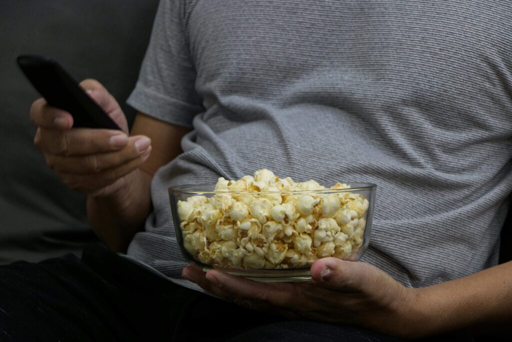 A man holding a bowl of popcorn, smiling as he enjoys watching television.

