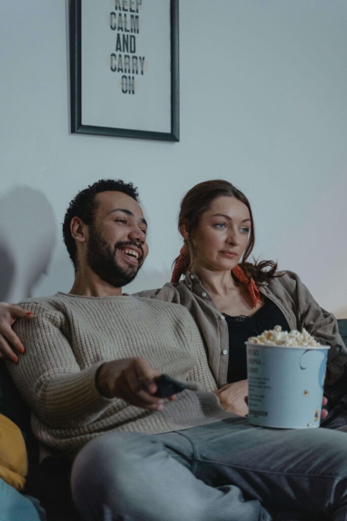 Smiling man and his partner watching TV together with a bowl of popcorn, under a wall art that reads “Keep Calm and Carry On.”