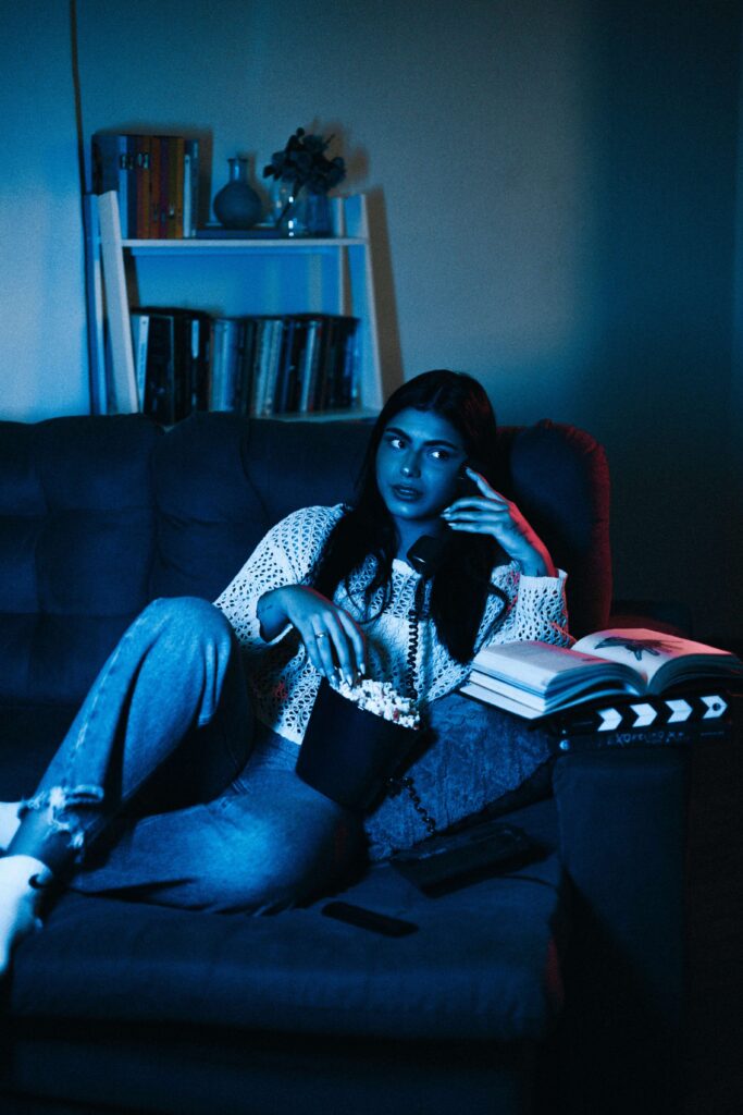 A girl talking on the phone, with a book in front of her, watching a movie and eating popcorn.